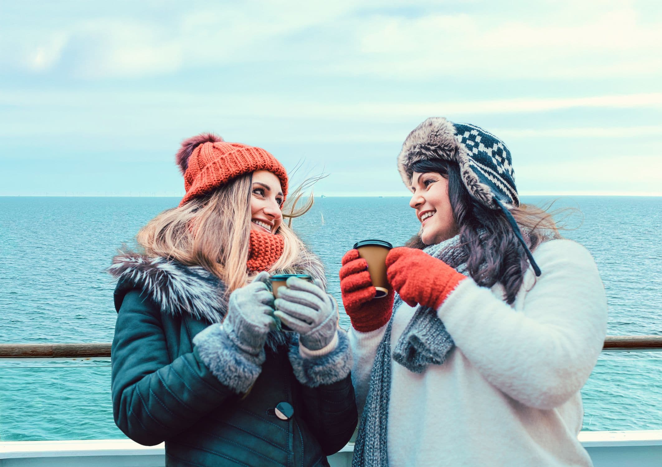 To veninder med huer på står på Scandlines' færgedæk med kaffekrus i hånden. 